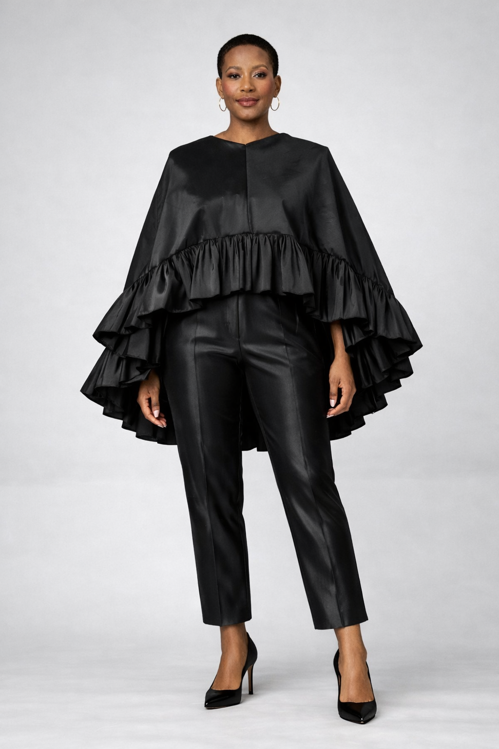 Woman wearing an elegant black silk taffeta two-piece set featuring a flowing ruffle cape and tailored pull-on pants.